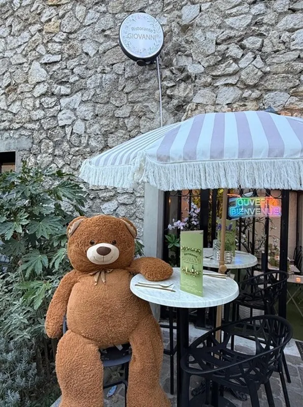 Le grand ours en peluche assis à une table de la terrasse, sous le parasol rayé